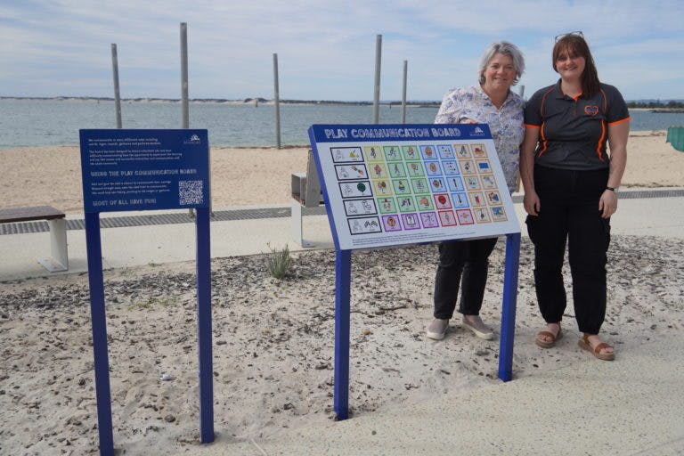 Two women stand beside the communication chat board sign, erected alongside a concrete path. In the background is Koombana Bay beach.