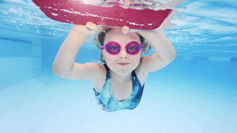 Child diving underwater with red kickboard and pink goggles